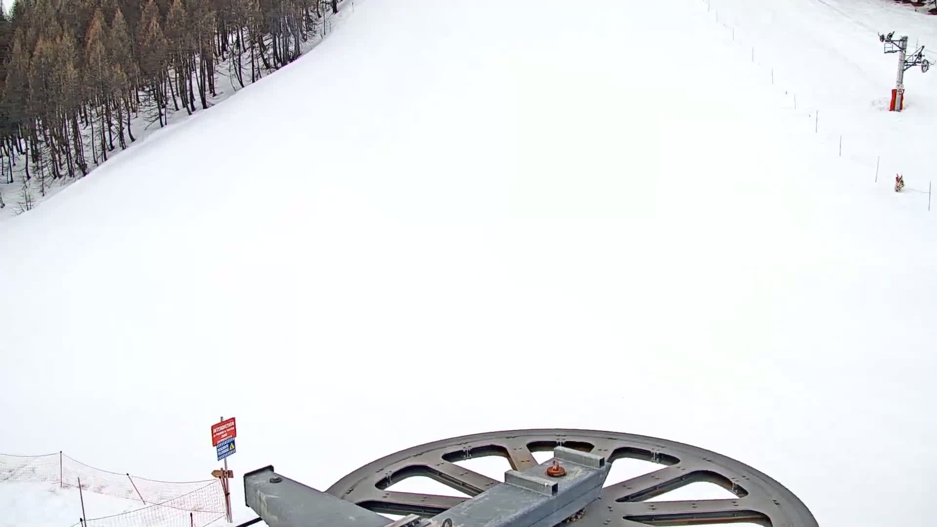 A large metal wheel of a ski lift is visible in the foreground, overlooking a snow-covered ski slope with a line of trees and another ski lift in the distance on a bright, snowy day.