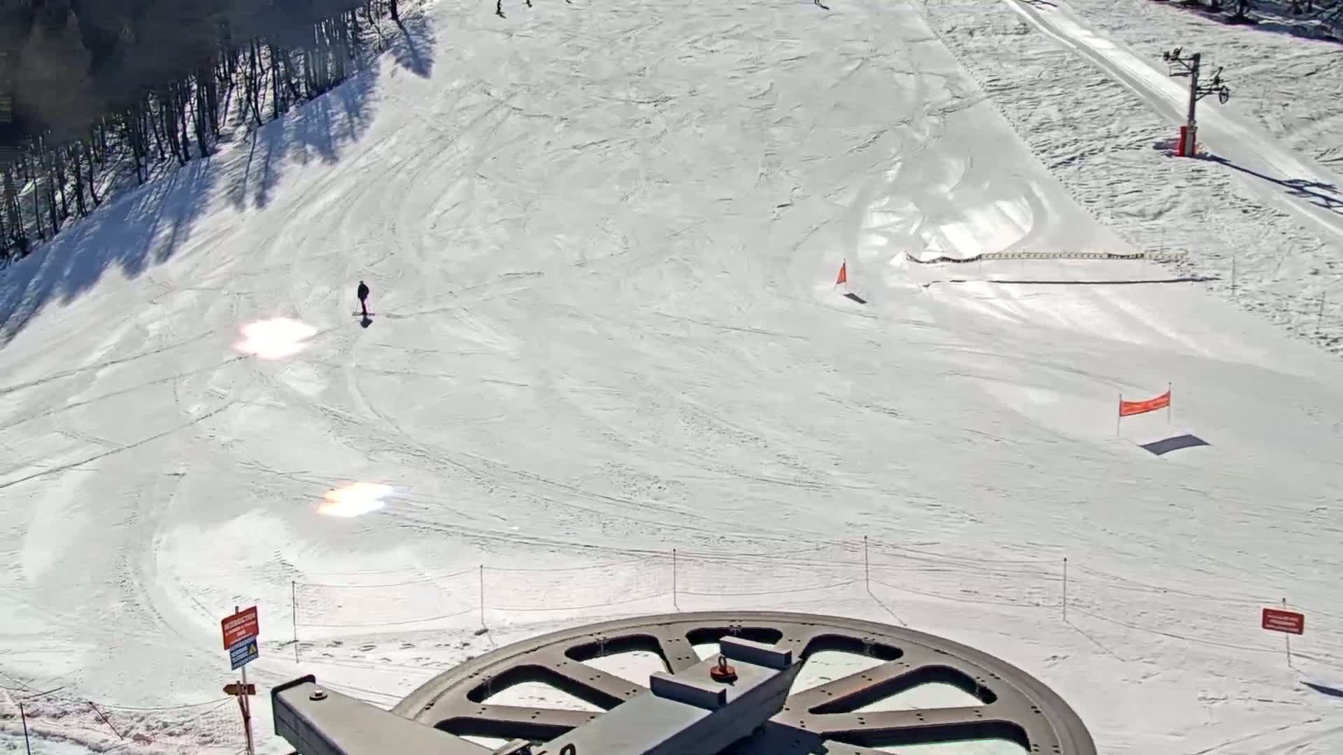 A snowy ski slope with a lone skier, a ski lift, and a large pulley wheel in the foreground on a sunny day.