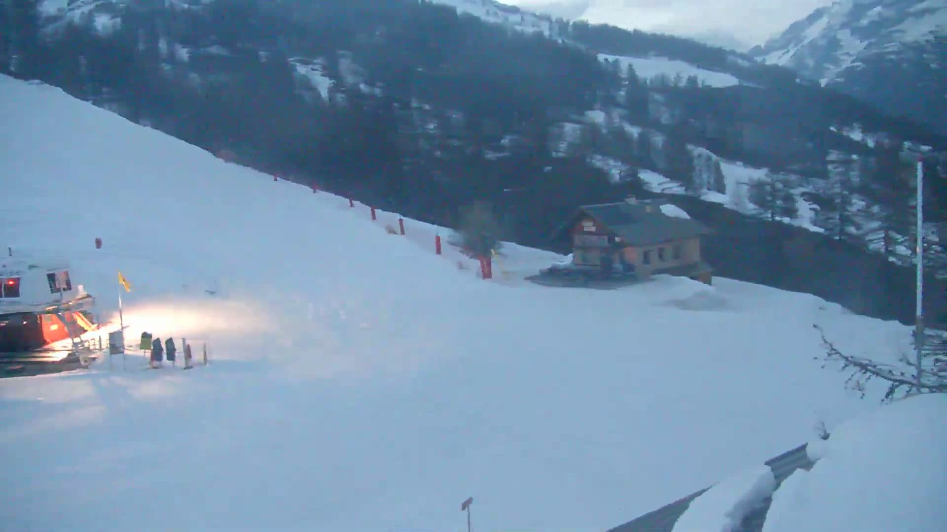 A snow-covered ski slope with a small building and a snowcat at the bottom, situated on a mountainside under overcast skies.