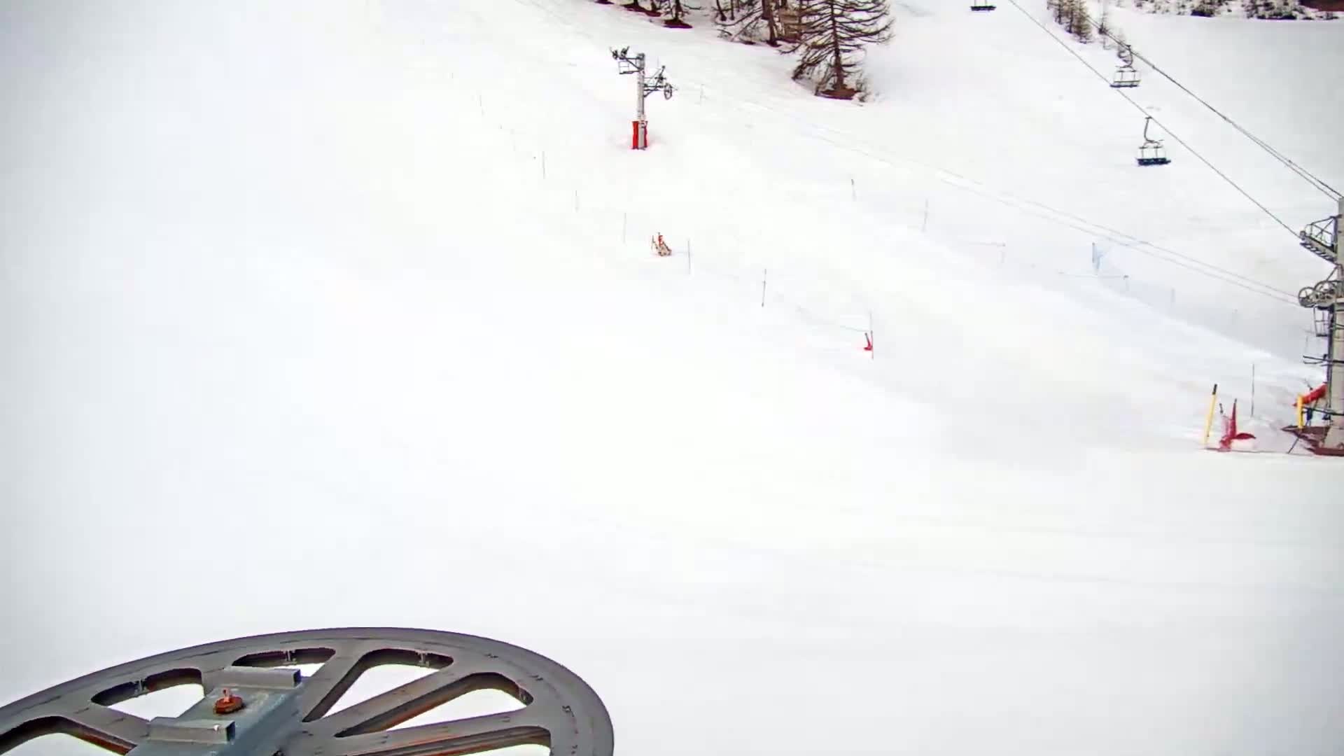 A snow-covered ski slope is viewed from a chairlift, showing ski lifts and markers in the distance.