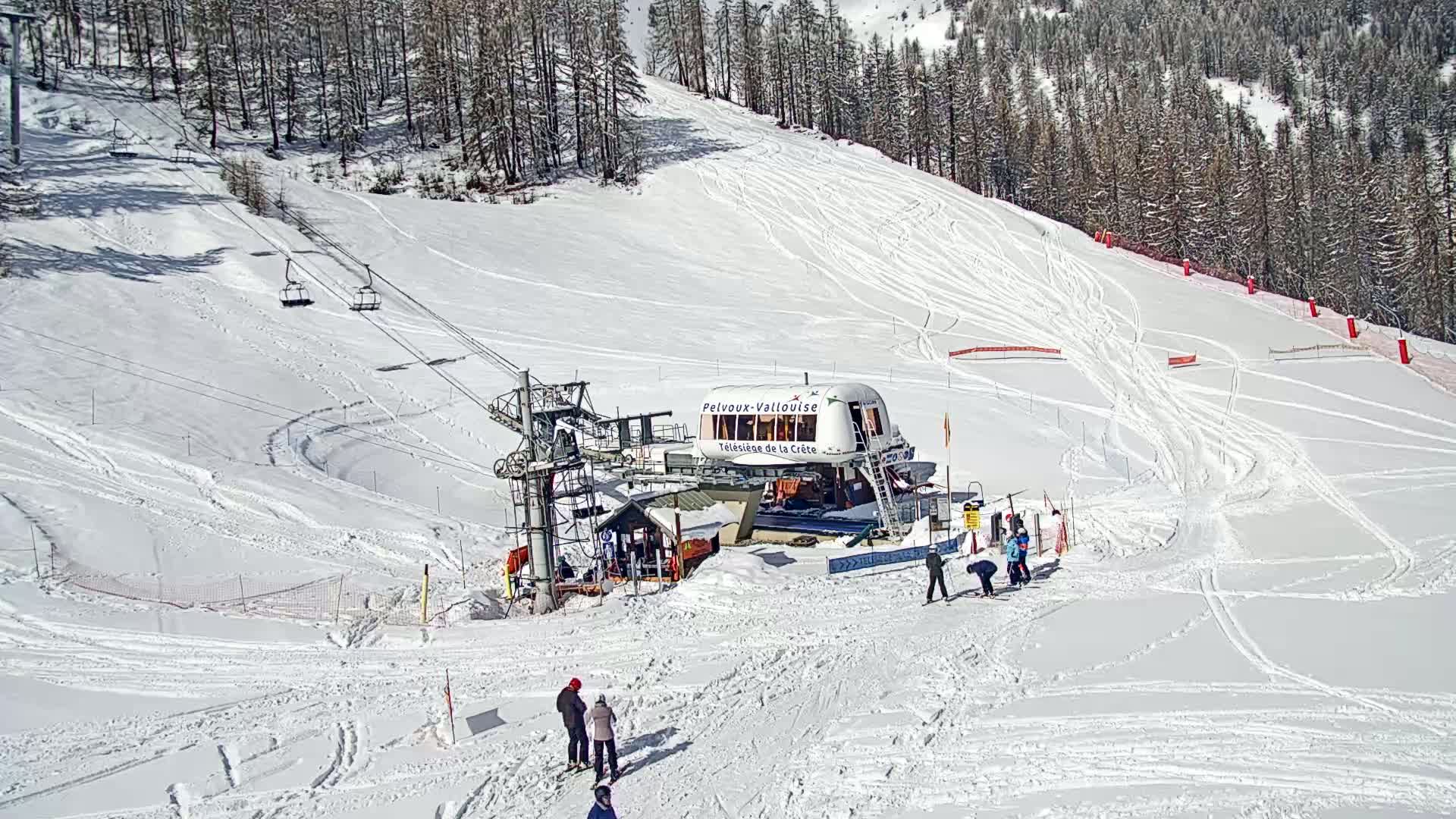 A ski lift station sits at the bottom of a snow-covered ski slope on a sunny day, with skiers gathered near it and several ski tracks visible on the slope.