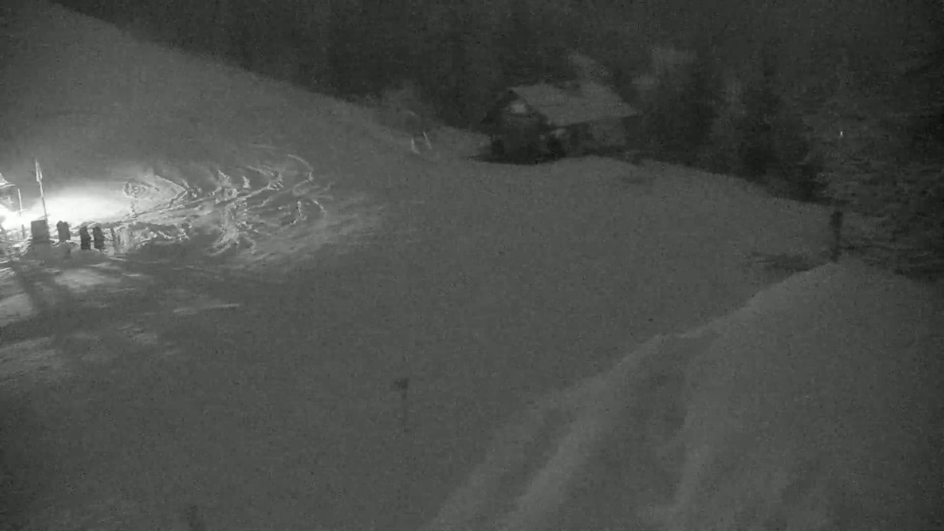 A snow-covered landscape at night shows a small building and tire tracks in the snow.