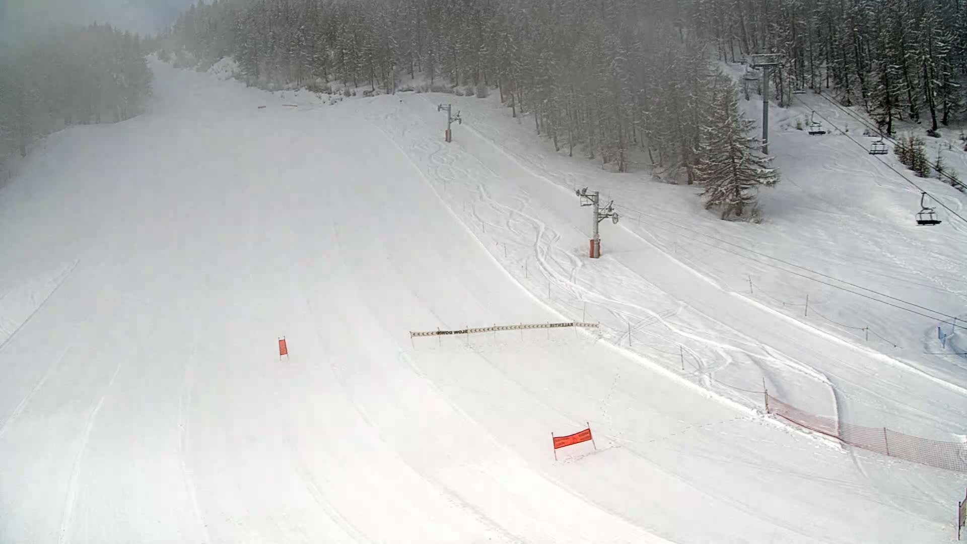 A snow-covered ski slope with several ski lifts and markings is visible under foggy conditions.