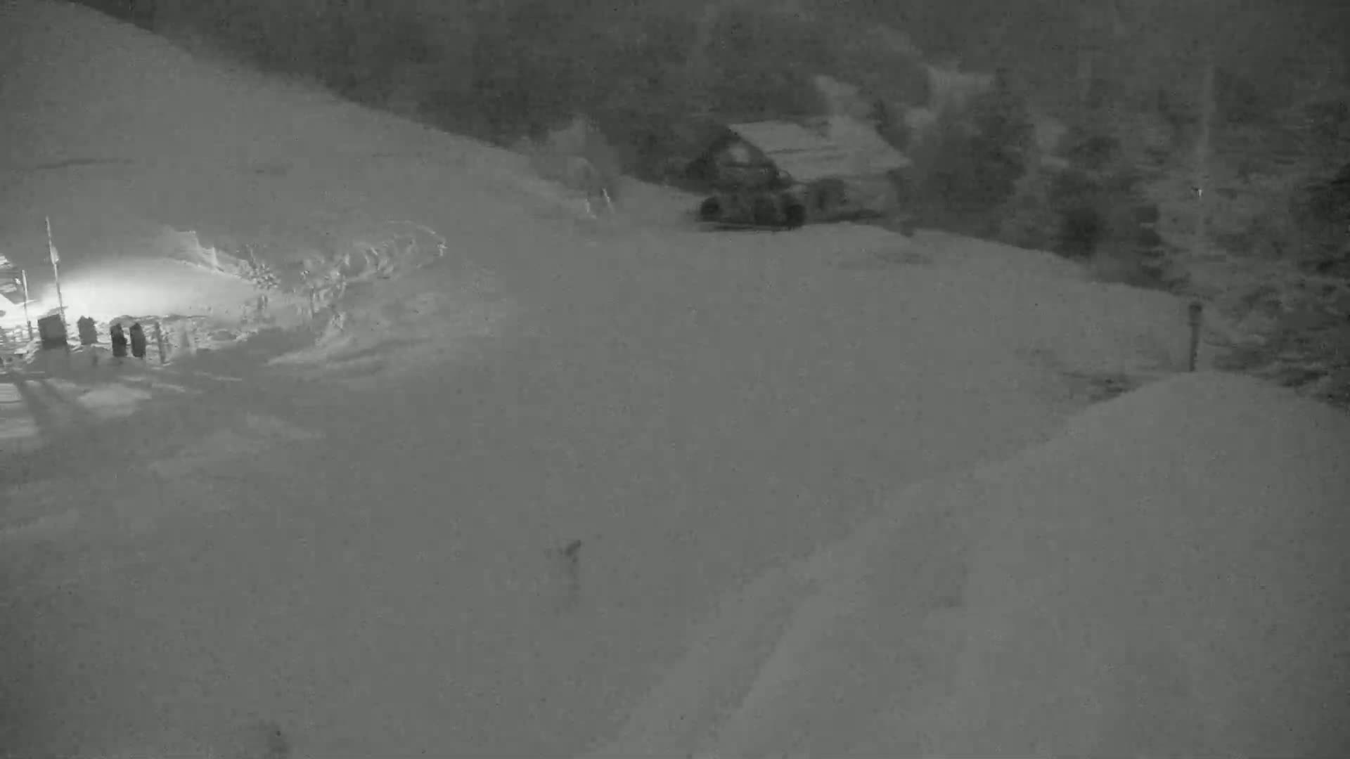 A snow-covered slope at night, with a small building and a lone figure visible in the distance under dark, snowy conditions.