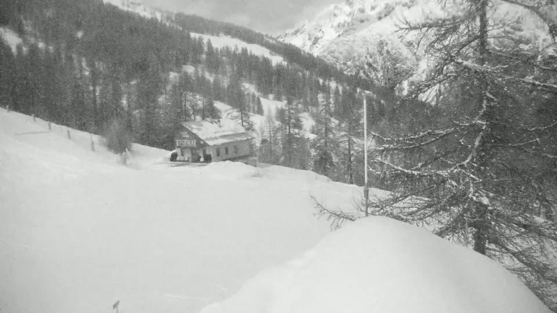 A snow-covered mountain landscape features a small building nestled among snow-laden evergreen trees under an overcast sky.