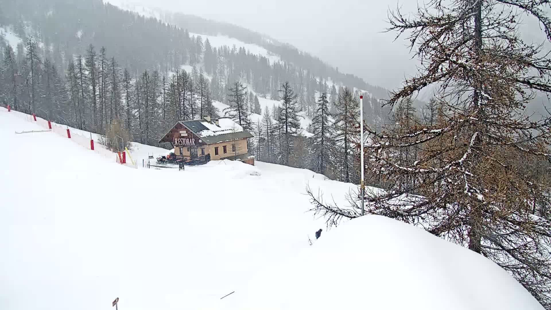 A snow-covered ski slope with a small building and sparse trees, under a heavy, overcast sky with falling snow.