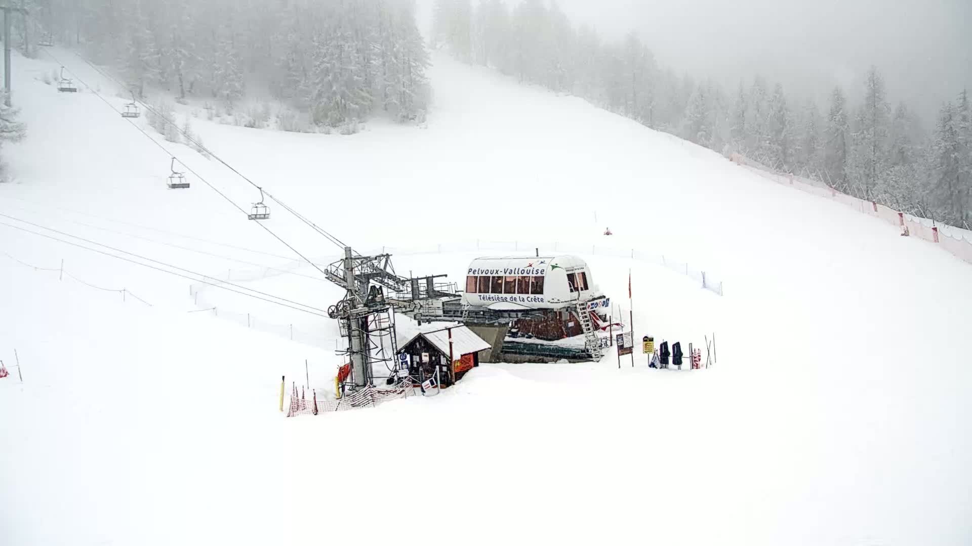 A ski lift station sits atop a snow-covered hill in foggy conditions, with several empty chairlifts extending down the slope.