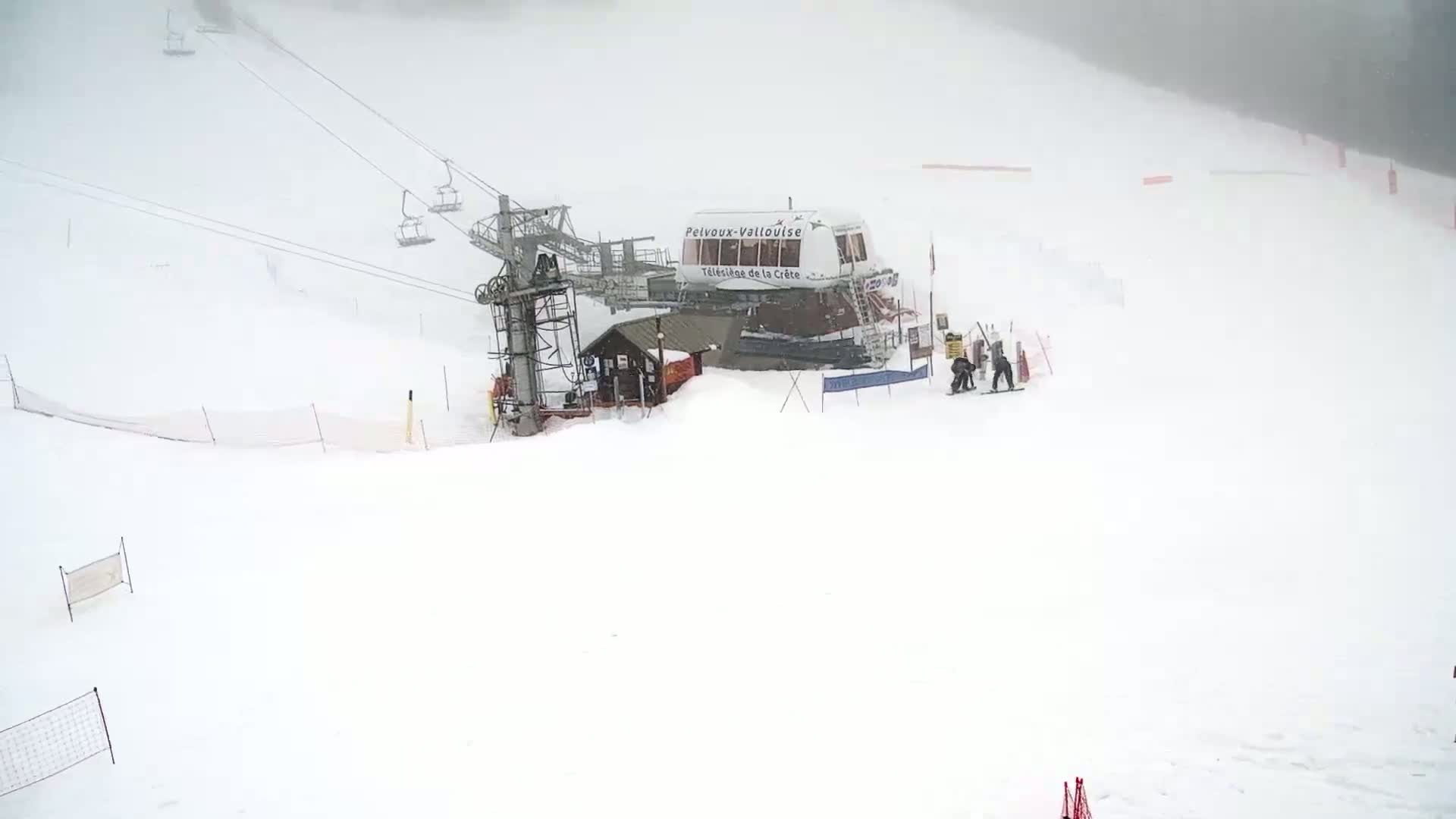 A ski lift and a few skiers are visible in a snowy landscape on an overcast day.