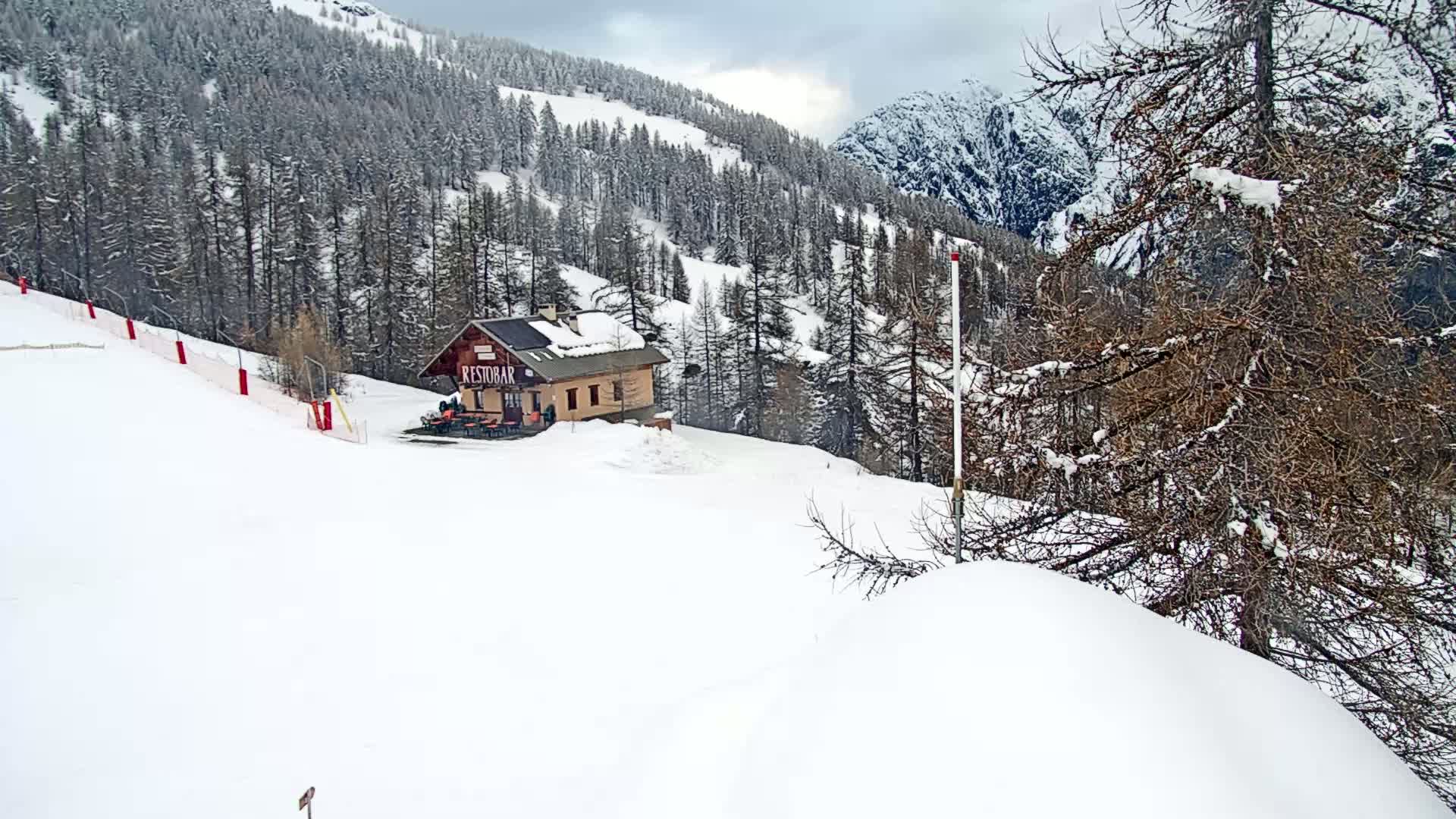 A snow-covered ski slope with a small building and snow-laden trees under an overcast sky.