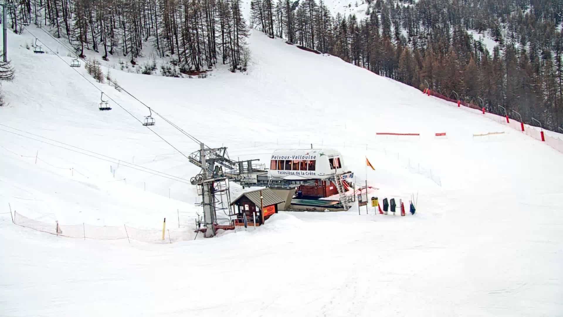 A ski lift base station sits at the bottom of a snow-covered ski slope surrounded by a coniferous forest under a cloudy sky.