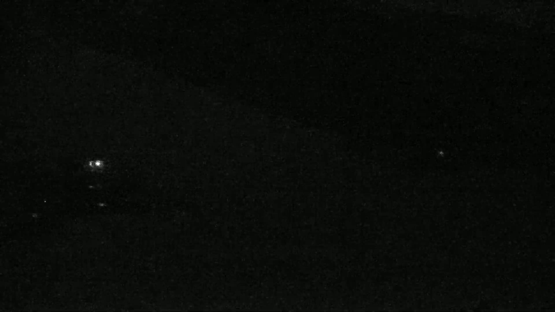 A nighttime, low-light image shows a few faint lights in the distance, possibly from vehicles or buildings, against a dark, mostly clear sky.
