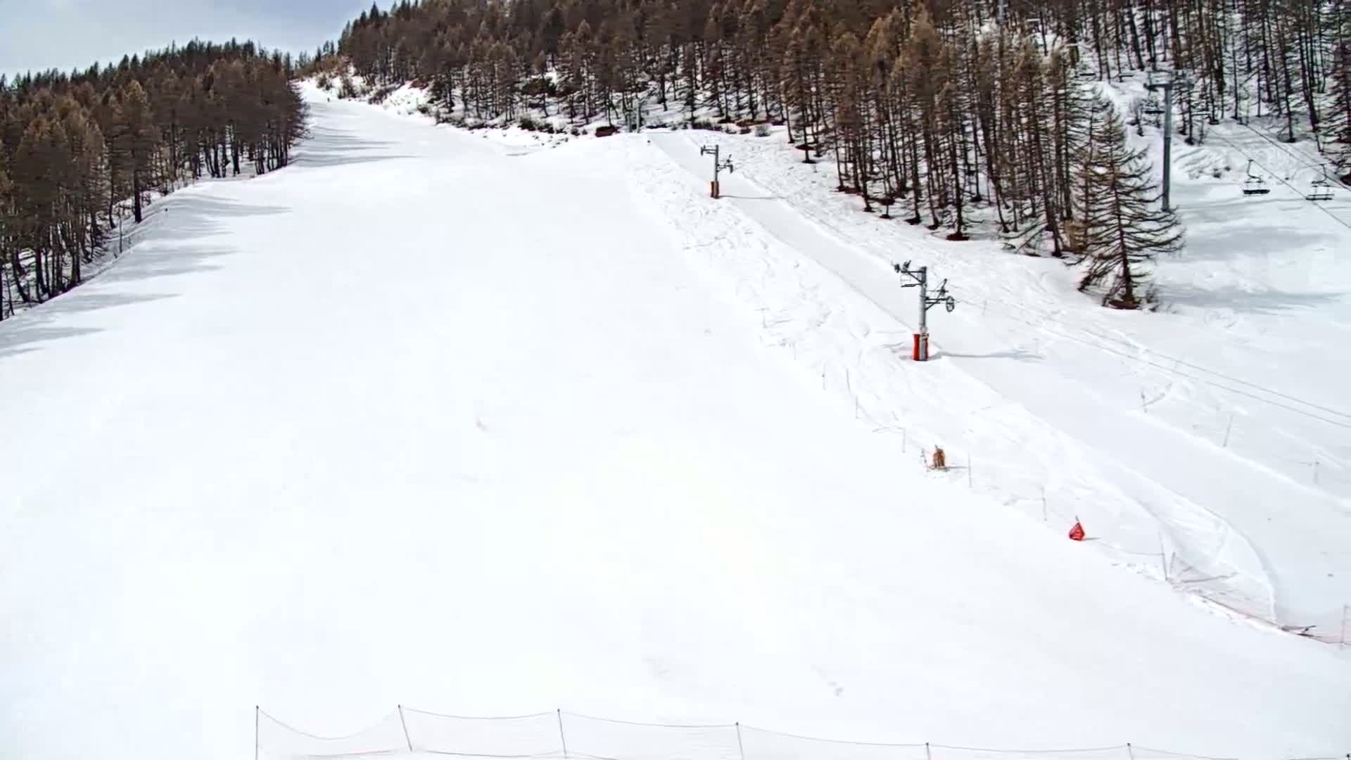 A snow-covered ski slope with two chairlifts is visible beneath a mostly cloudy sky, bordered by a dark evergreen forest.