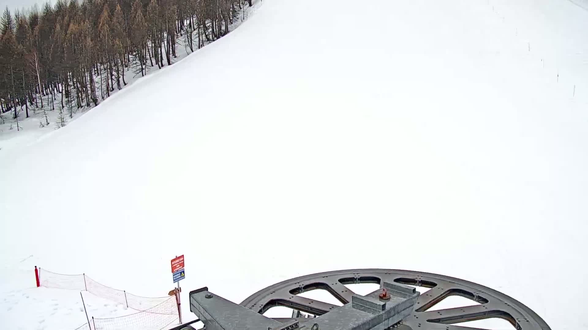A large metal wheel is situated on a snowy hillside next to a dark forest under bright, overcast conditions.