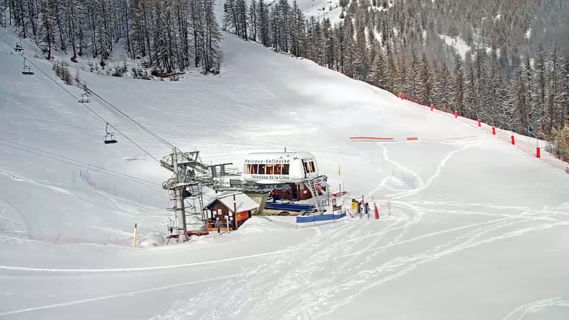 A ski lift with several empty chairs ascends a snow-covered slope bordered by a pine forest on a bright, snowy day.