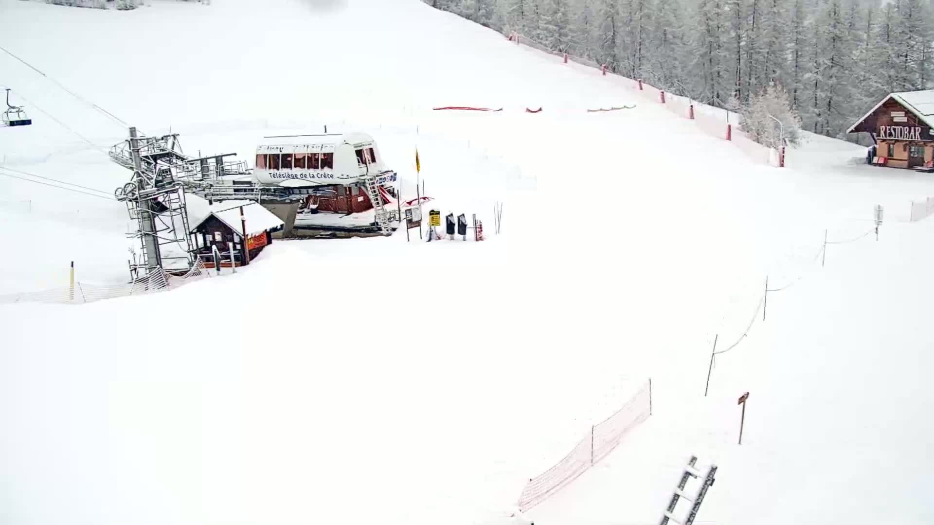 A snow-covered ski slope features a chairlift, a small building, and a snow-covered wooded area in the background during a snowy day.