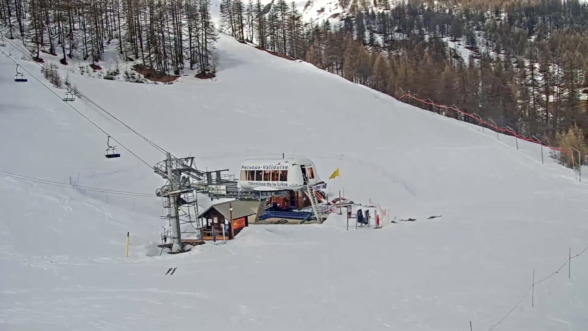 A chairlift and its base station sit on a snow-covered ski slope, backed by a coniferous forest under a clear sky.