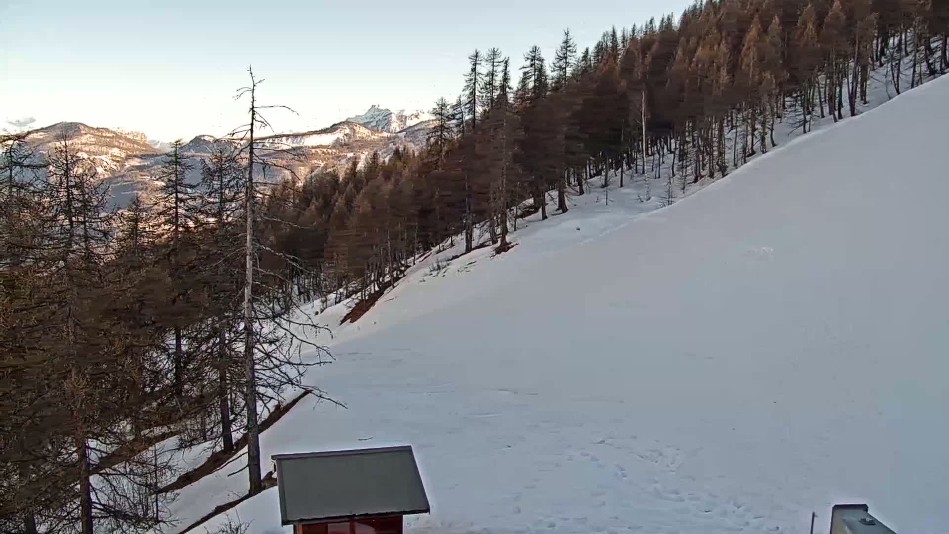 A snow-covered hillside slopes down from a dark pine forest toward a small building, with mountains in the background under a clear sky.