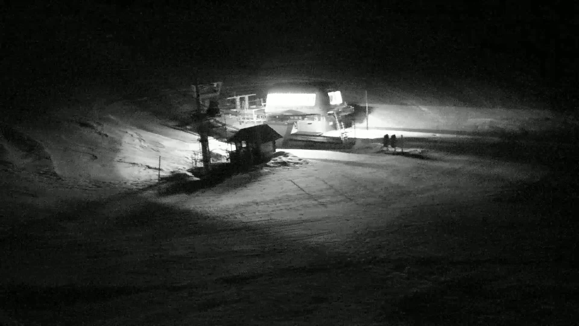 A snow-covered ski slope at night is illuminated by artificial light, showing a small building and two figures near a snowcat.