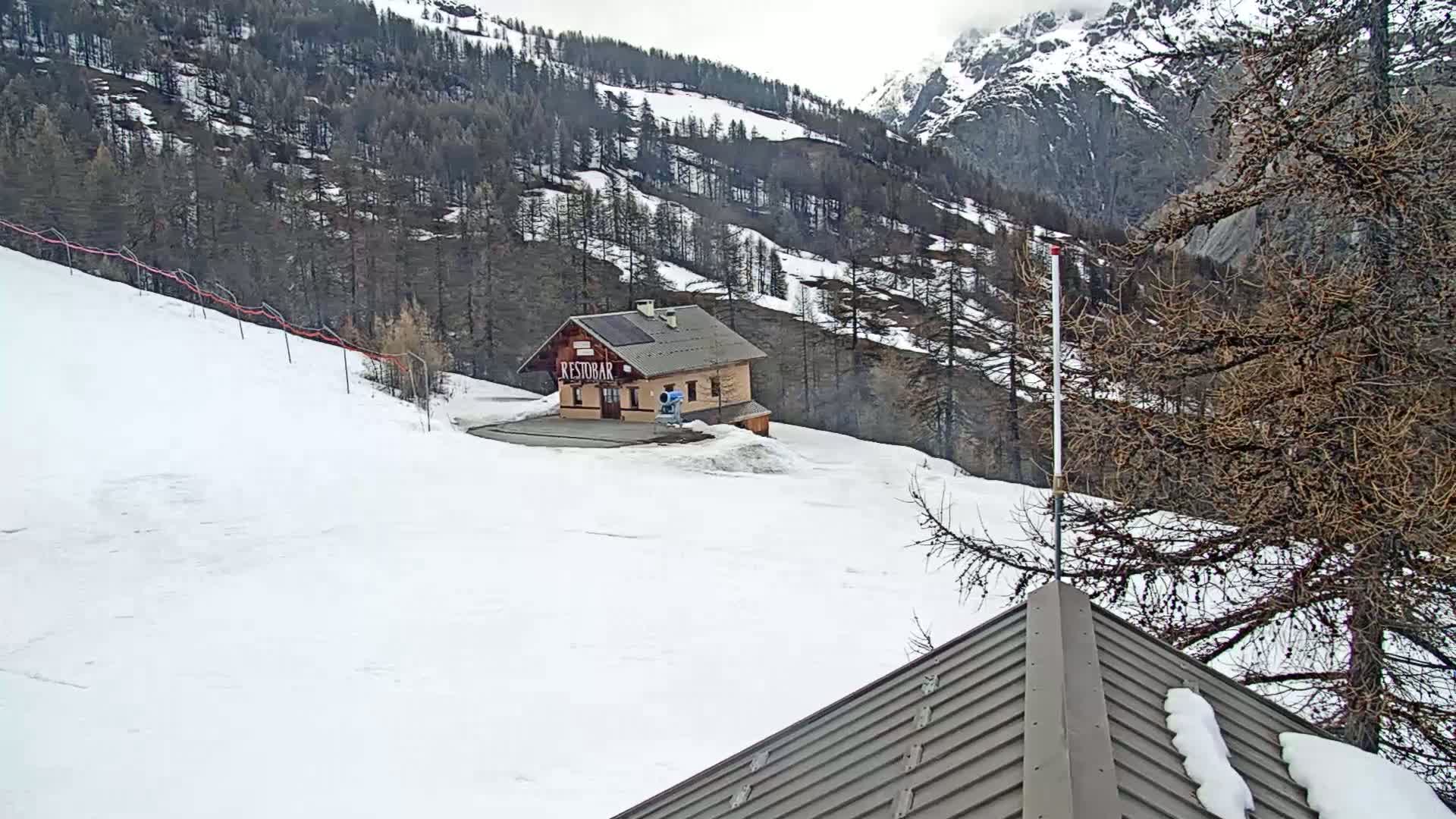 A snow-covered mountain slope with a small building and snowmaking equipment, under an overcast sky.