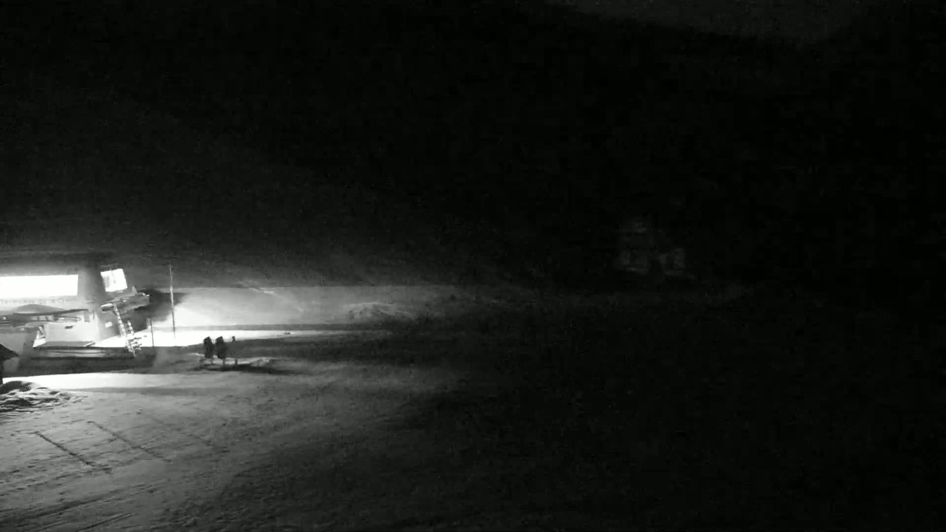 At night, two figures stand near a lit building in a snow-covered area.