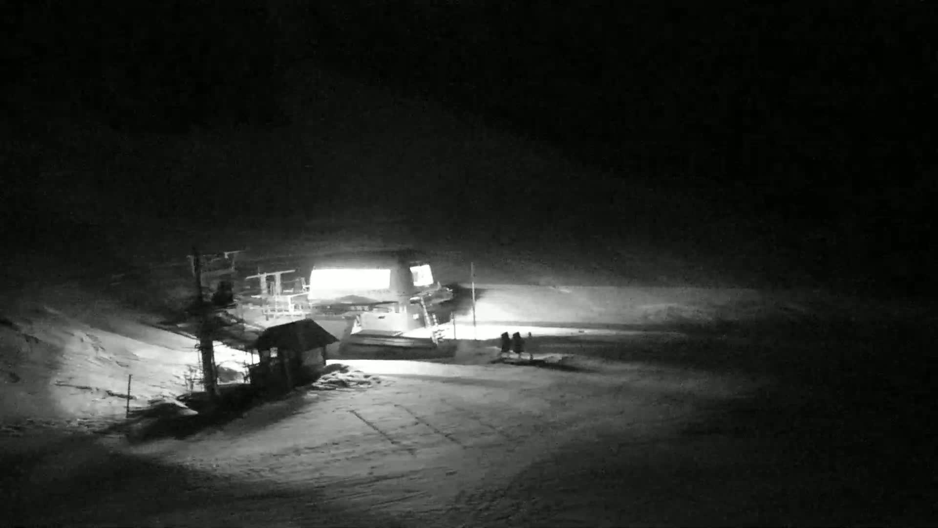 A snow-covered ski slope at night, illuminated by the lights of a lift station and a small building.