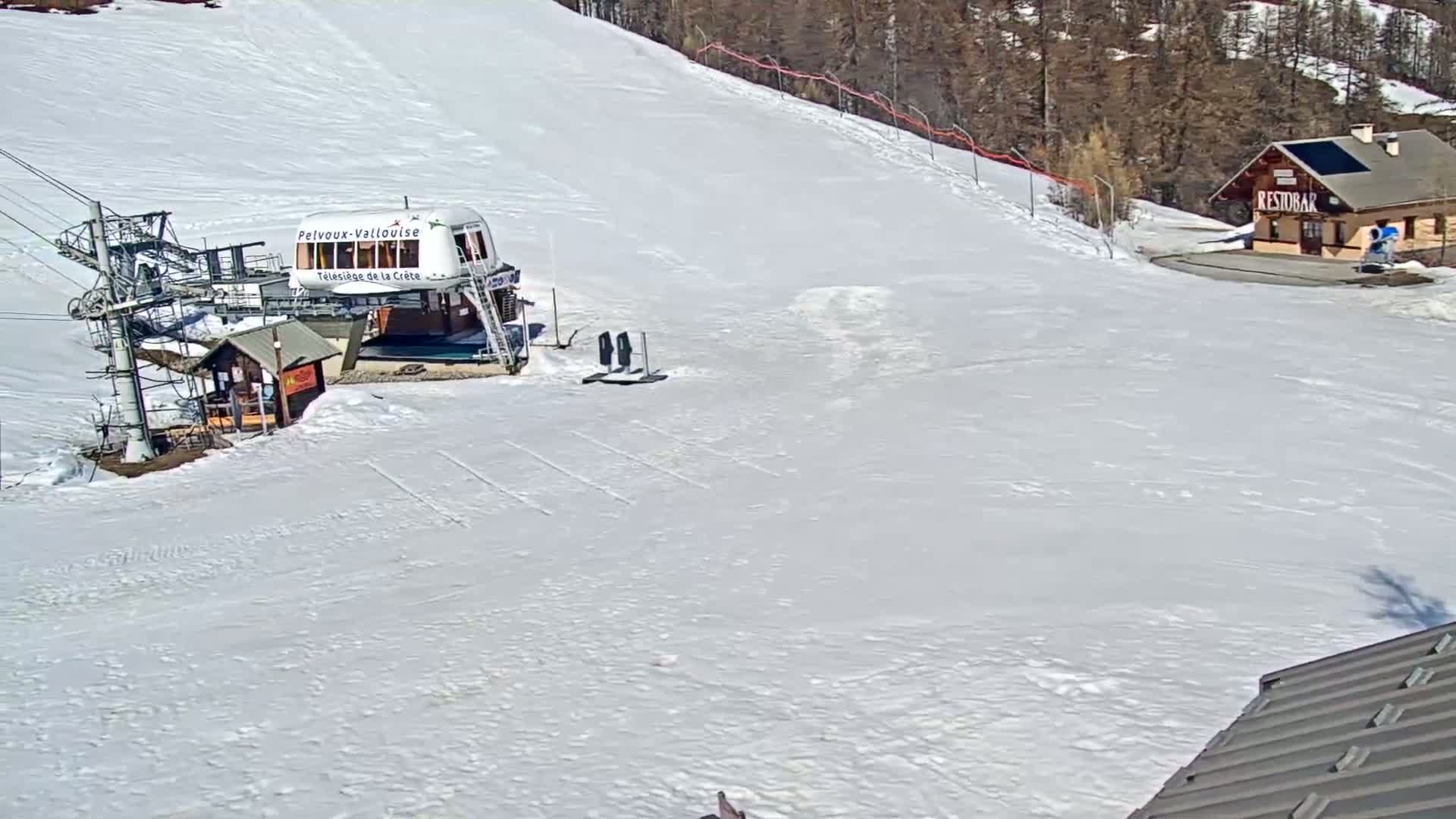 A chairlift station and a small building sit on a snow-covered ski slope under a sunny sky.