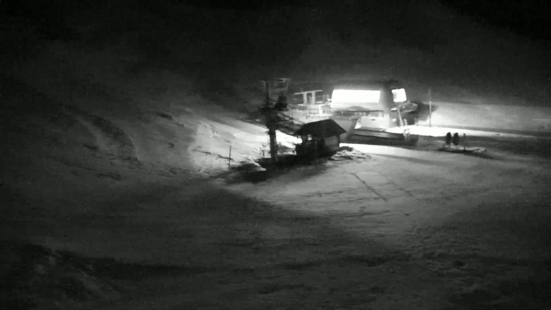 A snow-covered ski slope at night, illuminated by artificial lights, shows a ski lift and a small building.