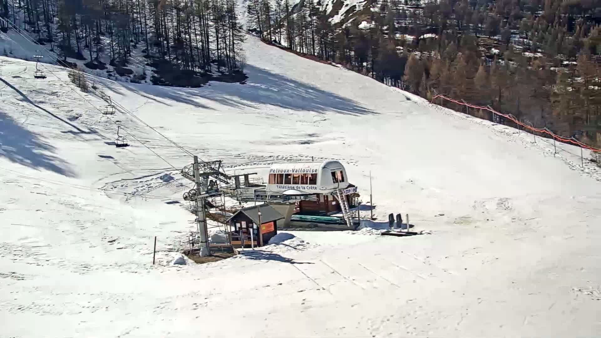 A ski lift and its terminal building are situated on a snow-covered slope on a sunny day, surrounded by leafless trees.