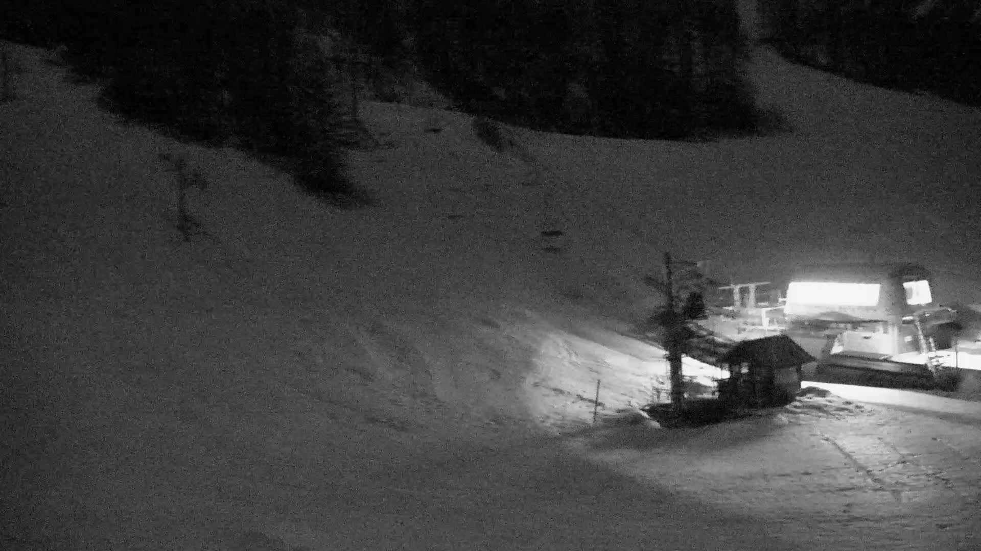 A snow-covered slope at night is illuminated, showing a small building and ski lift equipment near a larger structure.