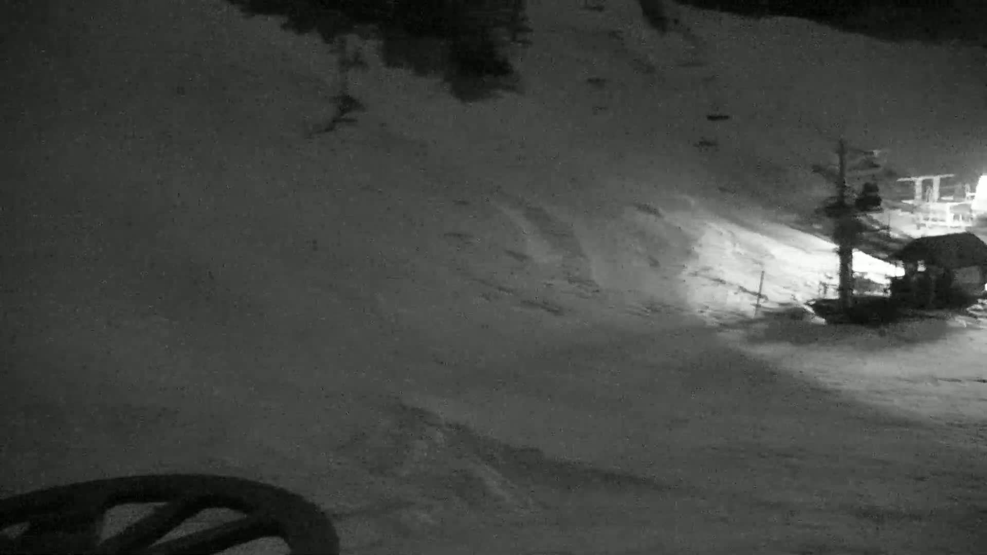 A snow-covered slope at night is illuminated by artificial light, showing a small structure near a ski lift.