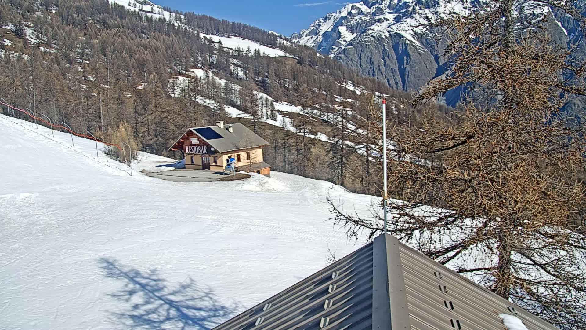 A snow-covered mountain slope with a small building and bare trees under a sunny sky.