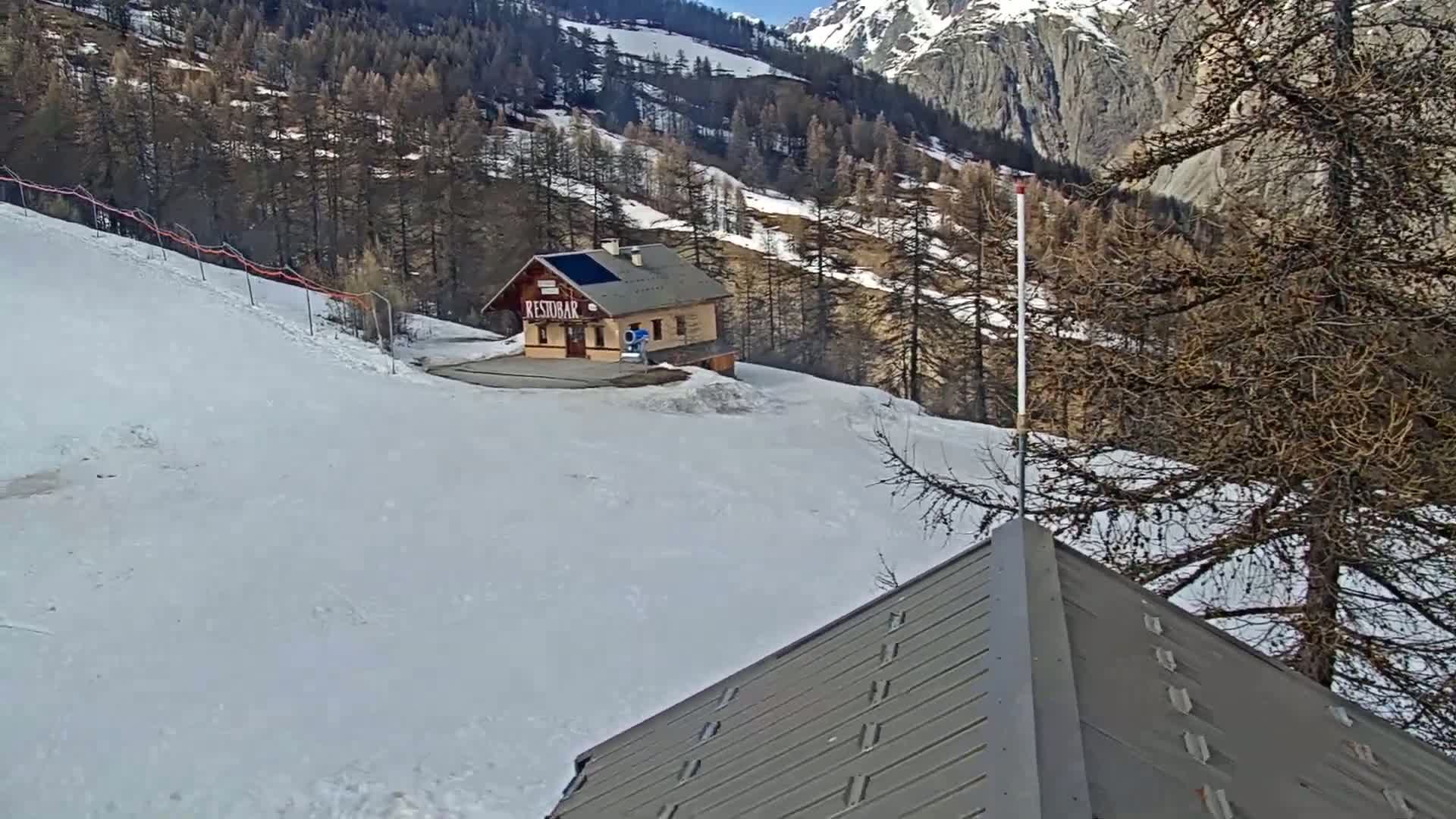 A snow-covered mountain slope with a small building and a section of a gray roof in the foreground under a sunny sky.