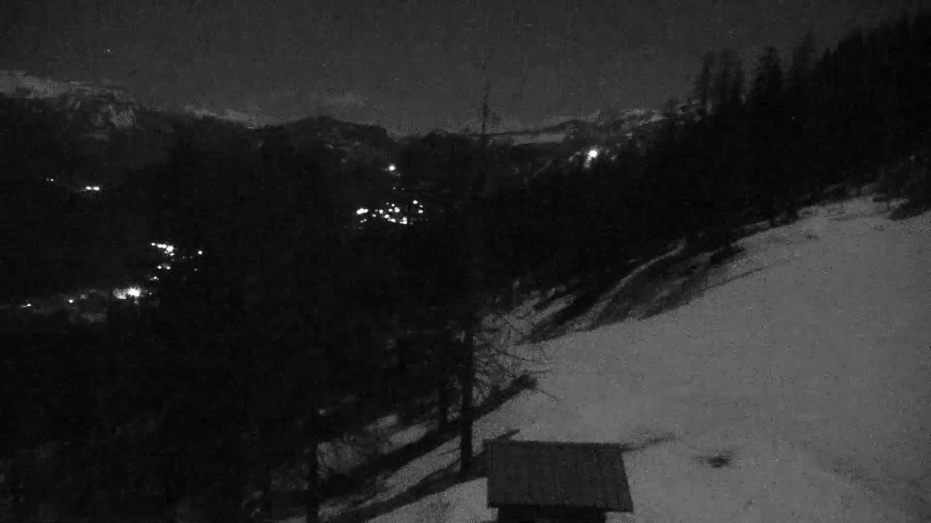 A nighttime view of a snow-covered slope with a small structure in the foreground, overlooking a dark valley with distant lights.