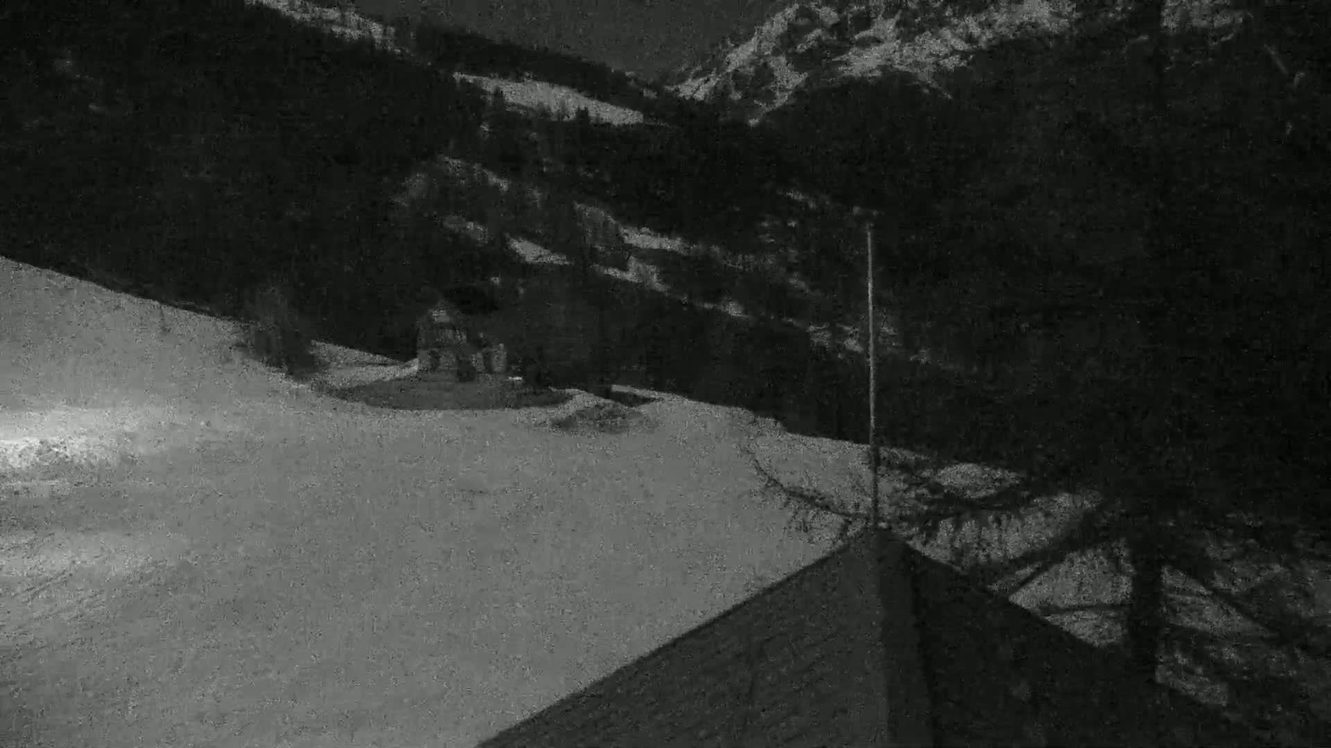 A nighttime, snow-covered hillside slopes down to a small building, with dark trees and a mountain in the background.