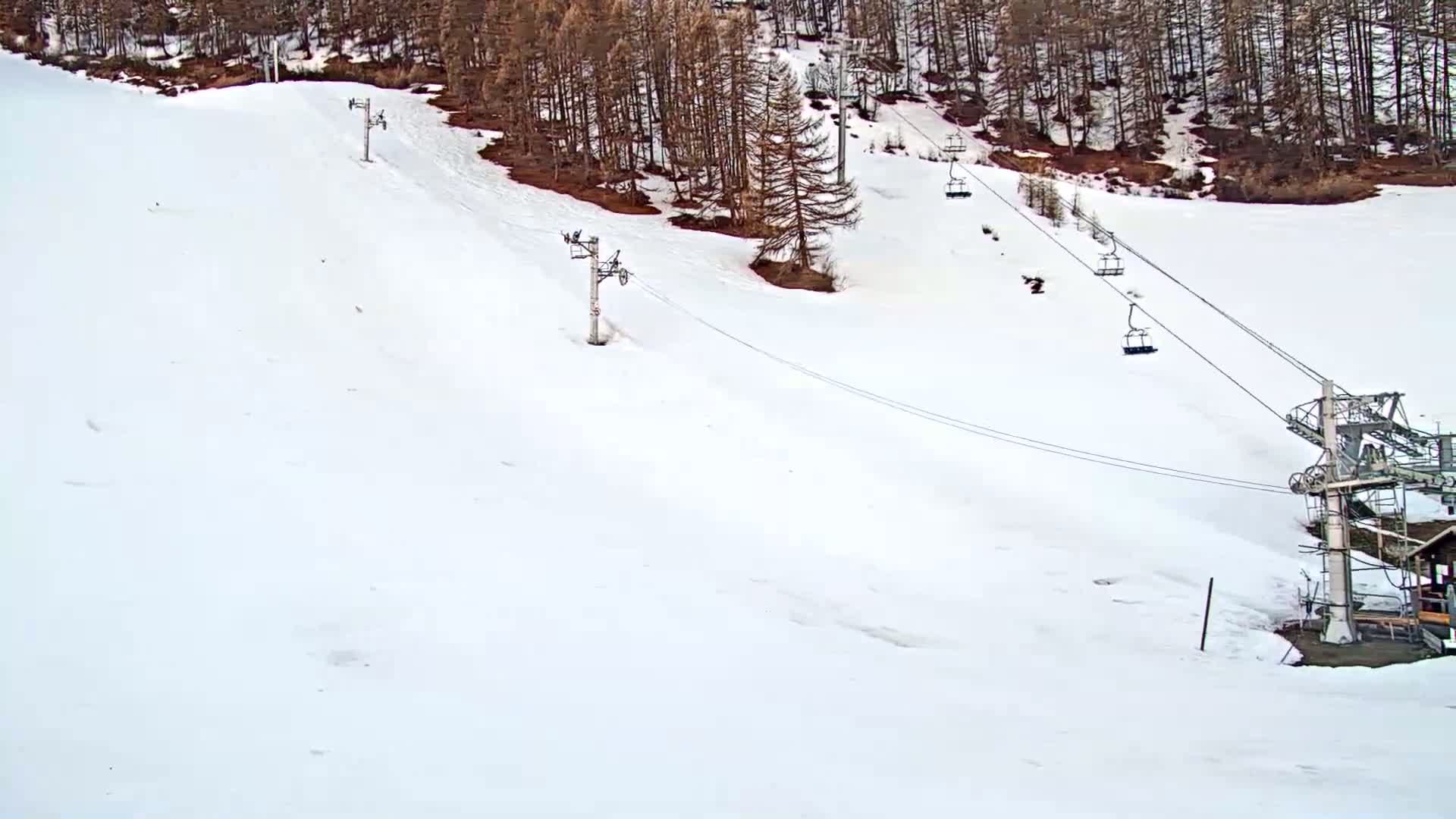 A snowy ski slope with several chairlifts and a few skiers or snowboarders visible, bordered by a dark forest.
