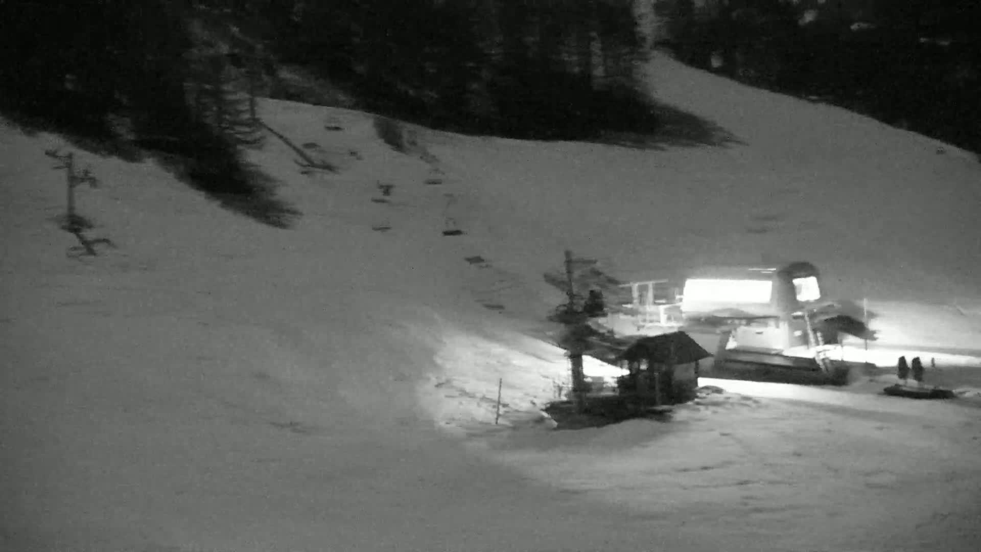 A snow-covered ski slope at night is illuminated, showing a ski lift, a small building, and a snow groomer.