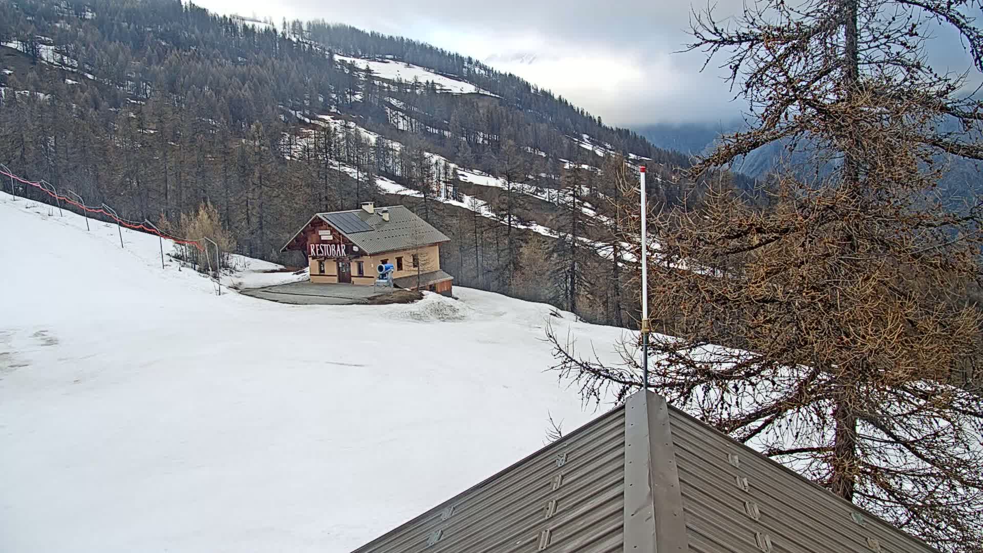 A snow-covered slope with a small building and bare trees under a cloudy sky.