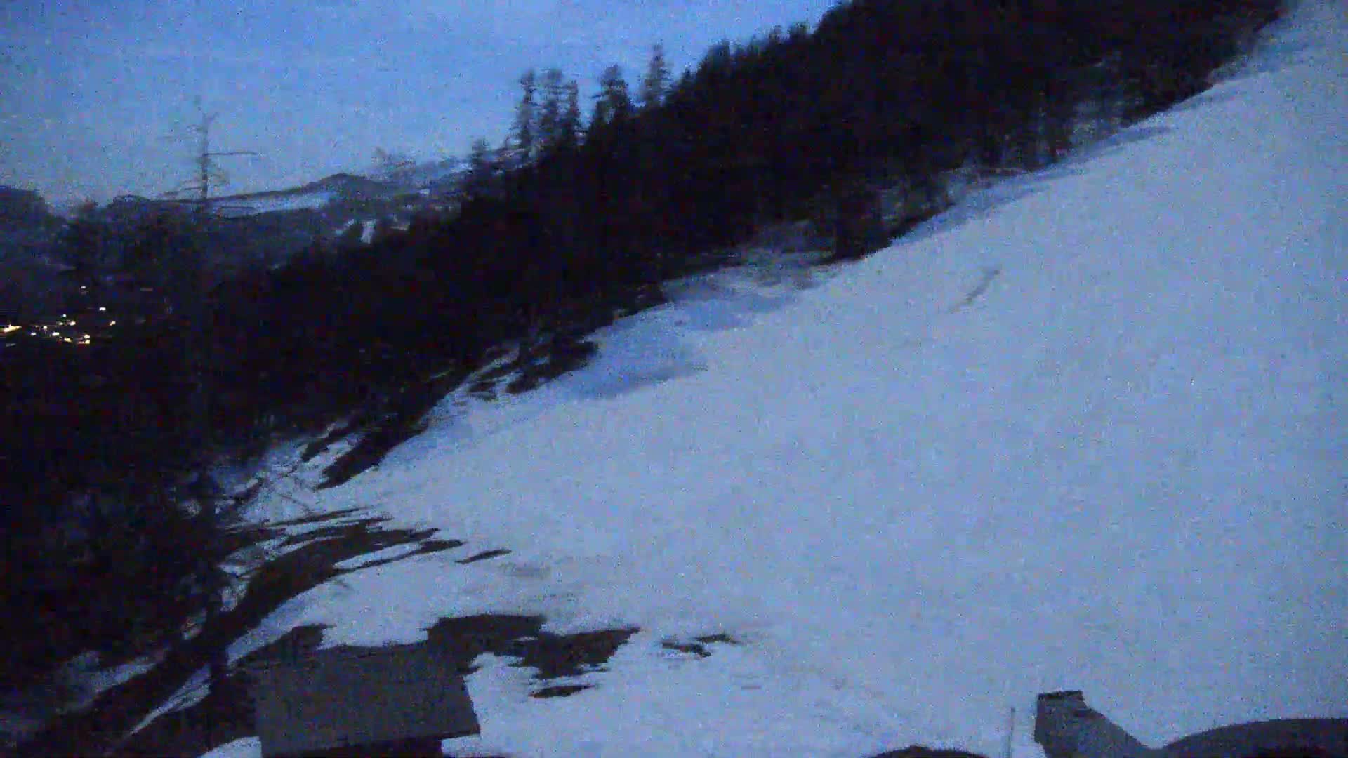 A snow-covered hillside at dusk, with a dark forest in the background and a few lights visible in the distance.