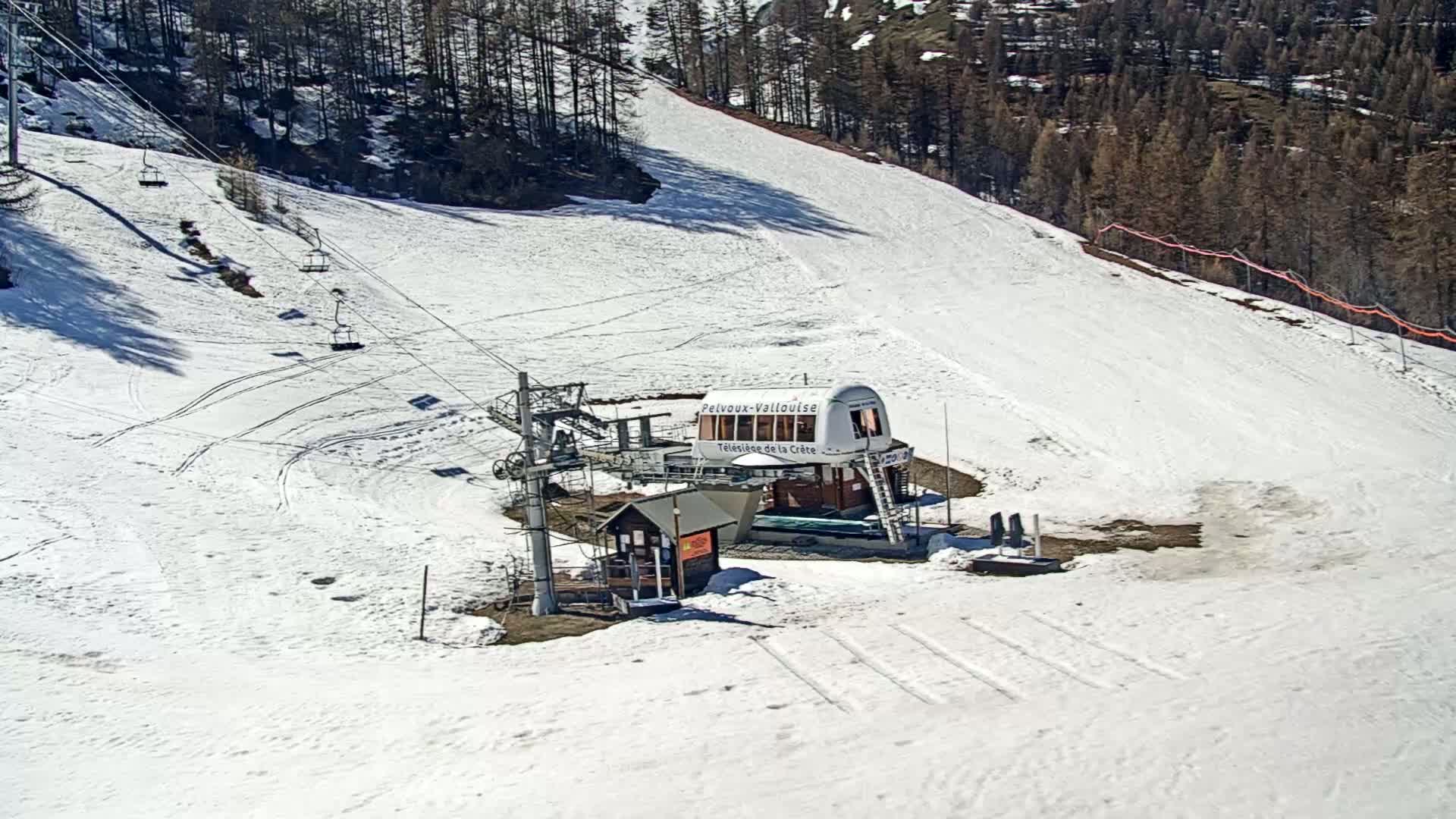 A ski lift station with a cable car and several chairs sits on a snow-covered ski slope, surrounded by a coniferous forest under sunny conditions.