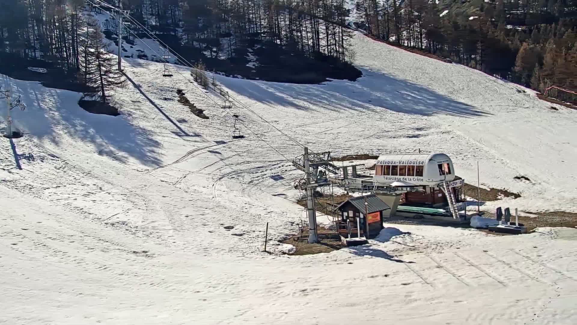 A ski lift and a small building sit on a snow-covered slope under a sunny sky, surrounded by dark trees.