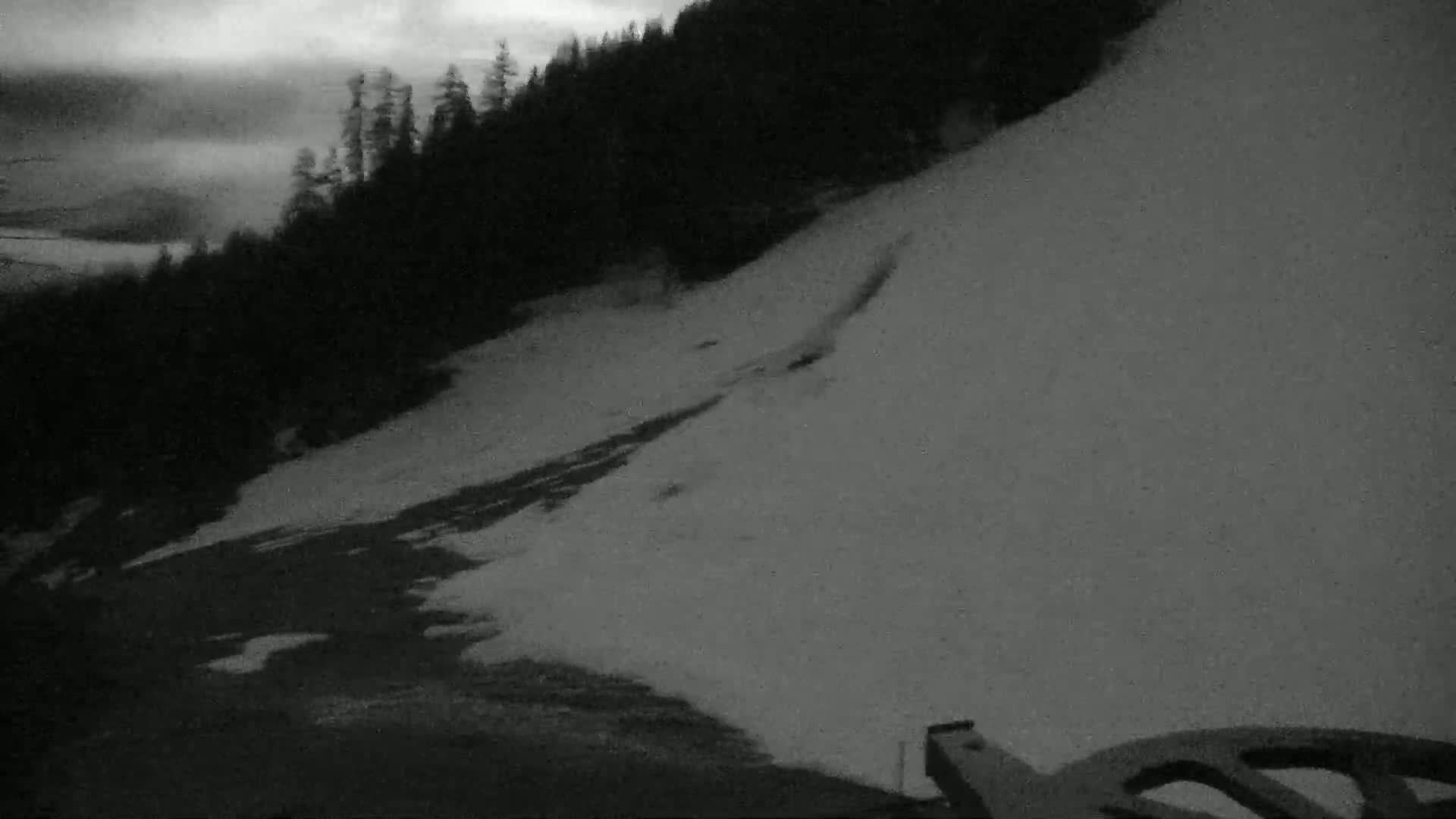 A snow-covered hillside at dusk, with a dark forest in the background under a cloudy sky.