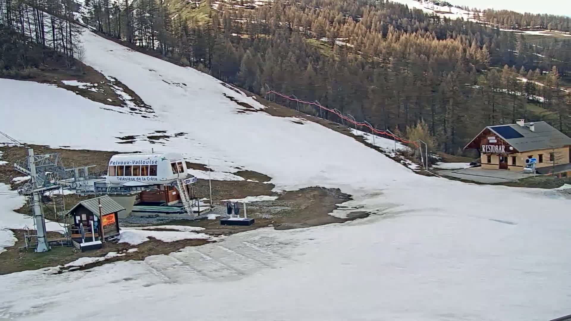 A ski lift station with patchy snow cover sits below a sparsely snow-covered hill and a forest, under a mostly clear sky.