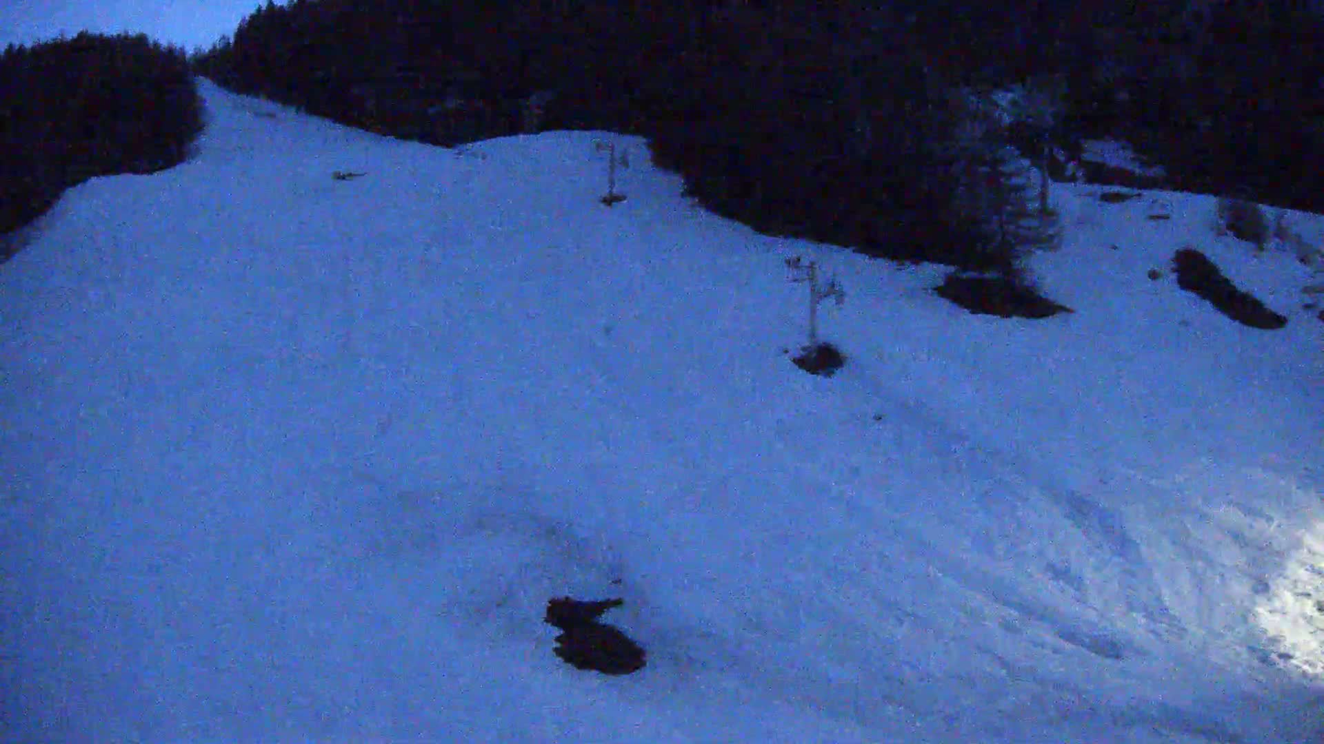 A snow-covered hill at night, with a few ski lift poles visible, is illuminated by a dim light source.