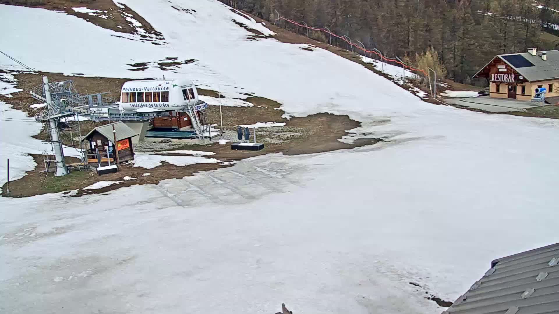 A ski lift station with a small building and melting snow sits below a mostly snow-covered hill.