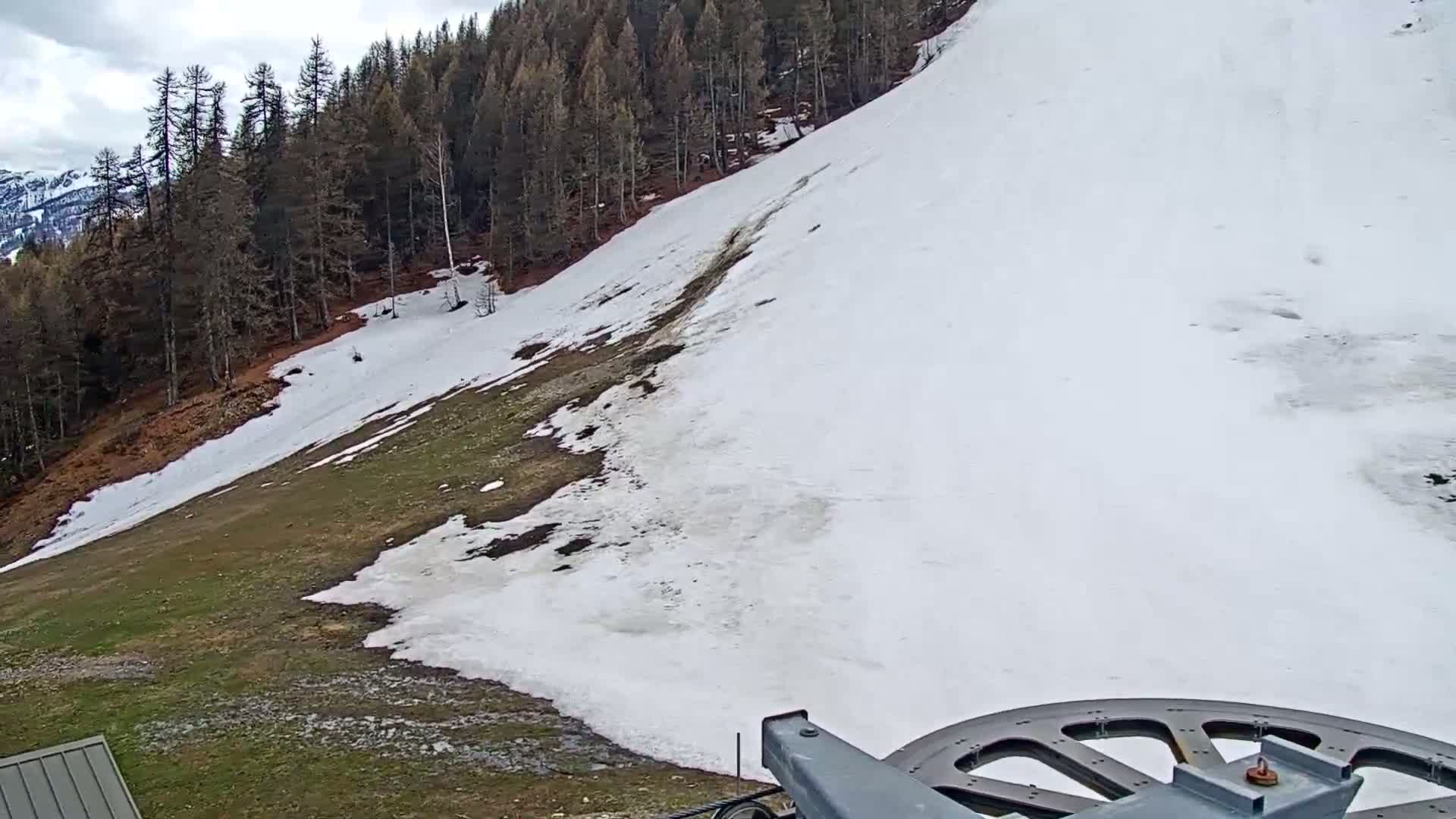 A snow-covered hillside slopes down towards a grassy area, partially melted snow remains on the ground, and a section of a ski lift is visible in the bottom right corner under cloudy skies.