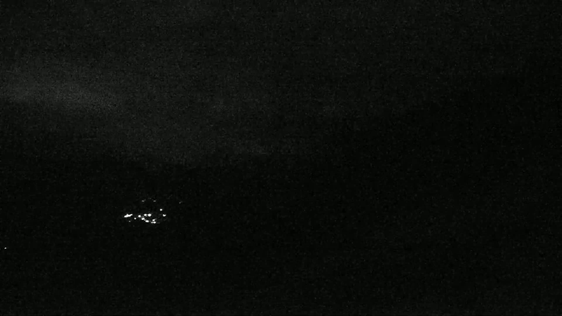A nighttime, low-light image shows a cluster of small, bright lights in the distance on a dark, possibly cloudy night.