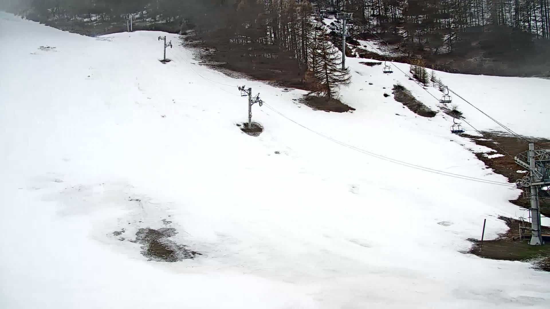 A snow-covered ski slope with several chairlift towers and dark green trees in the background, under overcast conditions.