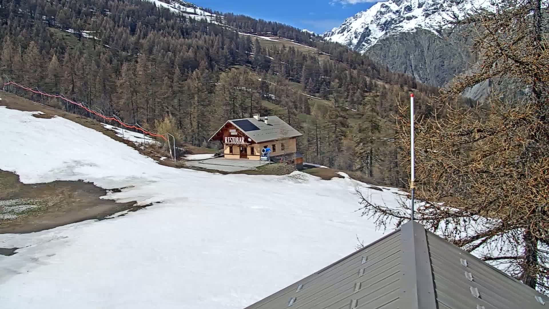 A small building sits on a snow-covered slope in the mountains on a sunny day.