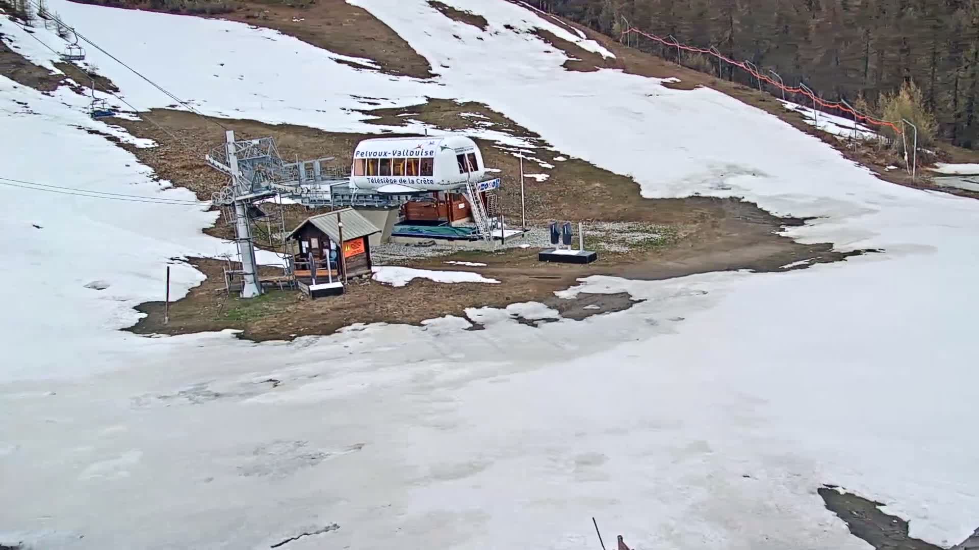 A ski lift station with a small building sits on a mostly snow-covered hill, with patches of bare ground visible.