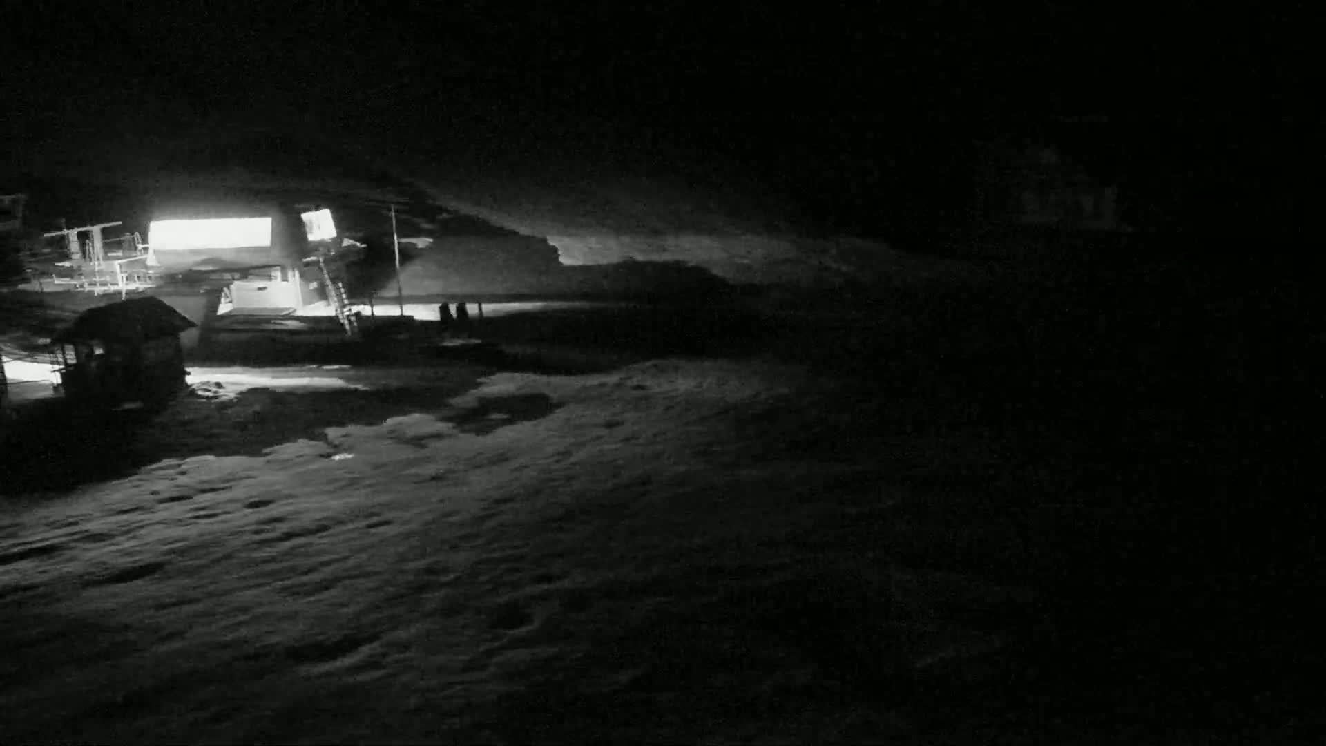 A nighttime, snow-covered scene shows a dimly lit building and several structures, possibly ski lifts, under a dark sky.